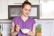 © sementsova321 - Photo of slim beautiful female model wears purple blouse, holds bowl with cereals, keeps to diet, eats healthy food. Young housewife prepares lunch for her child. Kitchen interior. Domestic concept