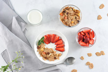 Plate With Corn Flakes With The Addition Of Fresh Strawberry Yogurt. Next To The Glass With Yogurt, Cups With Cereal And Strawberries. In The Frame, A Napkin, A Spoon. Light Background. Clos-up.