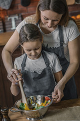 Canvas Print - Young girl and mother in aprons standing together and mixing vegetable salad on kitchen.