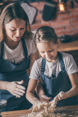 Poster - Young little girl and mother in aprons preparing the dough on kitchen table together. 