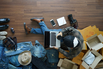 Top view of man holding coffee cup working laptop computer with fashion accessories on wooden floor from home. With postal parcel, Selling online ideas concept design