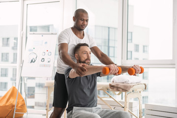 Wall Mural - Exercising equipment. Nice pleasant serious man sitting on the medball while holding dumbbells in front of him