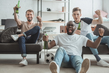 young happy multicultural male friends watching football match and celebrating with beer bottles in hands