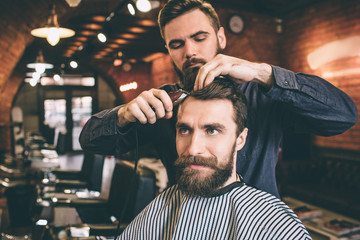 Hairstylist is using scissors for cutting hair. He is doing that very accurate. Te customer is looking straight forward and smiling.