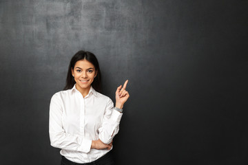 Sticker - Portrait of secretary or business woman with long dark hair wearing white shirt pointing finger upward on copy space, isolated over dark gray background