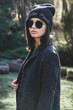 Street style portrait of a girl with black sunglasses and black hat on into the woods looking away