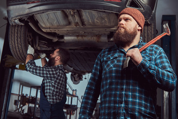 Two bearded brutal mechanics repair a car on a lift in the garage. 