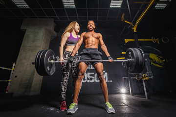 fit woman girl motivates african man while he doing deadlift with heavy barbell. man lifting barbell opposite window. emotional moment of lifting weight. affective lifting in gym. sport couple in gym.