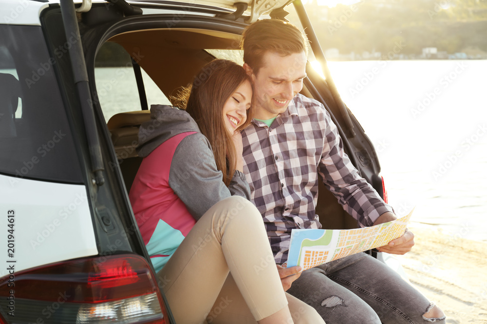 Beautiful young couple with map sitting in car trunk near river