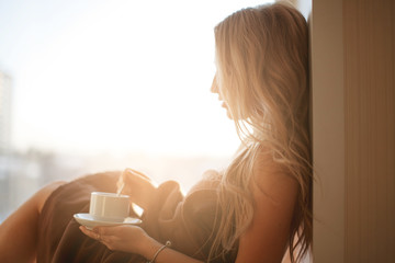 Young beautiful Girl drinking coffee sitting near the window in morning. Blond sexy naked female Boudoir photo standing in front of large window wrapped in blanket with her back to camera