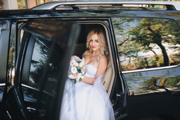 A beautiful bride with curly hair is sitting in the car and looking out the window. A nice bride in the city comes out of the car.