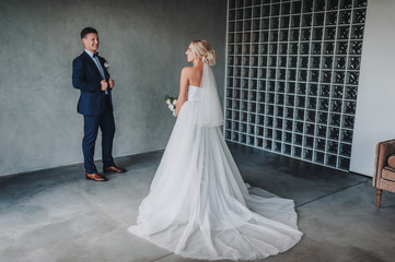 A stylish groom stands near a gray wall and looks at his beautiful bride in a long dress. Delicate and smiling newlyweds in a gray studio. Film effect.