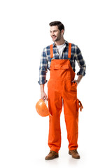 Wall Mural - Young builder in orange overall holding hard hat isolated on white