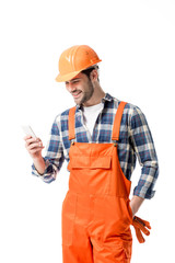 Wall Mural - Smiling handyman in orange overall and helmet using smartphone isolated on white