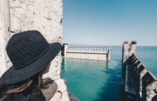 Girl With Hat Looking Through A Window Of Amazing Panorama Of Garda Lake In A Sunny Day From The Castle In Sirmione - Italy.