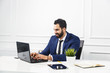 © proimagecontent - Bearded businessman dressed in blue suit work with laptop in white office interior