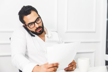 Wall Mural - Handsome bearded businessman wears white shirt and eyeglasses talk by phone in white office interior