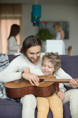 Sticker - Happy caring father and little son playing guitar sitting on sofa, loving dad holding music instrument having fun with kid boy, smiling daddy teaching child, family leisure activity at home, vertical