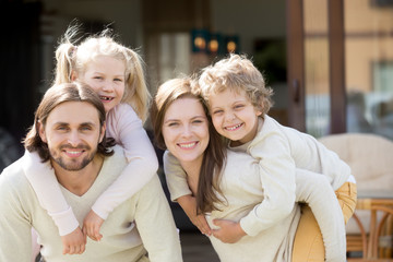 Wall Mural - Happy parents piggybacking son and daughter outdoors, smiling couple with cute children have fun together enjoy leisure time outside house looking at camera, active healthy family with kids portrait