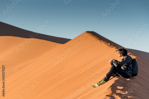 Zdjęcie XXL Młody męski podróżnika i fotografa obsiadanie na wierzchołku piasek diuna patrzeje wschód słońca lub zmierzch w pustyni Namibia, Afryka. Koncepcja fotografii podróży
