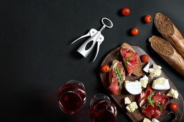 Wall Mural - Prosciutto, salami, baguette slices, tomatoes and nutson rustic wooden board, two glasses of red wine over black background