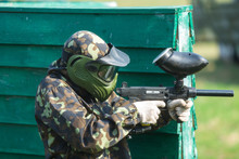 Boy In The Camouflage Holds A Paintball Gun  In One Hand And Protective Helmet , Standing On The Field With Group Of Players On The Background