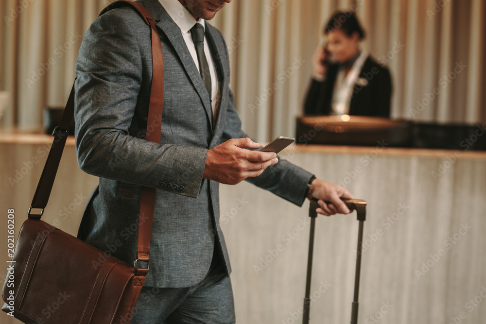 Businessman in hotel lobby with phone and luggage - obrazy, fototapety, plakaty 