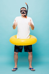 Full lenght Young handsome man with inflatable yellow ring and snorkel with thumbs up isolated on green background