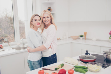 Wall Mural - Cheerful pretty mother and daughter hugging while cooking salad dinner having vegetables kitchenware dish frying pan on the table standing, girl getting experience from mother, leisure comfort concept