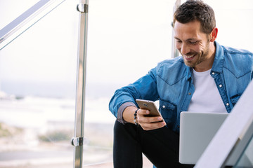 Handsome young smiling man sitting on a stair in the street working with a laptop and mobile phone. business concept
