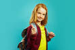 Positive schoolchild with backpack behind his shoulders shows his thumb, showing positive attitude to study, standing on blue isolated background. Red-haired girl goes to the first class.