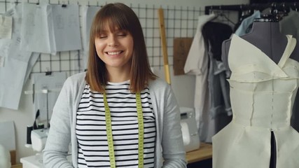 Wall Mural - Portrait of attractive young woman clothing designer with brown hair looking at camera. Smiling woman is standing beside dummy, light modern tailoring workshop in background.