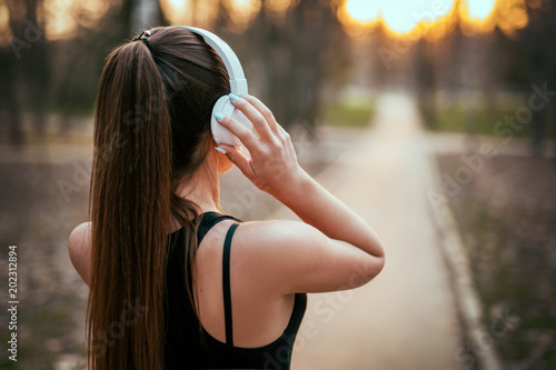 Fotomural sports girl standing with back in bluetooth headphones at sunset in the park