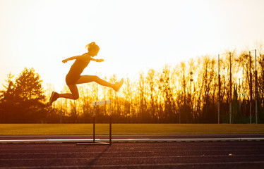 Canvas Print - Athletic woman jumping above the hurdle on stadium running track during sunset