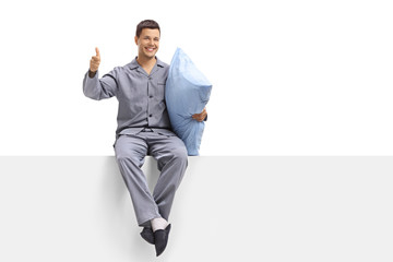 Poster - Young man in pajamas seated on a panel holding a pillow and making a thumb up sign