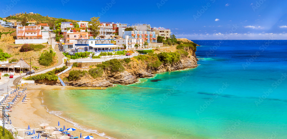 Sandy Varkotopos beach in sea bay of resort village Bali. Views of shore, washed by waves with sun loungers and parasols where sunbathing tourists. Greek houses stand on the rocky shore. Crete, Greece - obrazy, fototapety, plakaty 