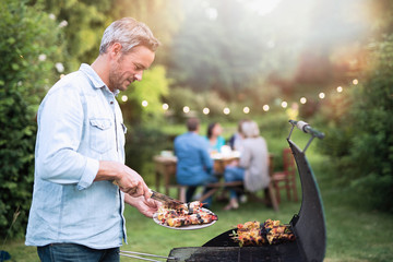 Wall Mural - in the summer,  a handsome man in his forties prepares a barbecue for his friends gathered around a table in the garden