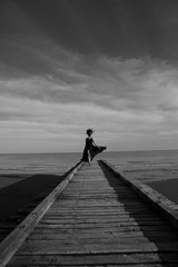 Beautiful woman in a dark luxury dress with big flower crown on head walking on a wooden pier. Dark clouds on background. Black and white