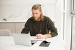 © Anton - Young man sitting at the table with laptop and making notes in notebook at home isolated