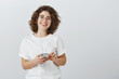 © Cookie Studio - Sister learns spanish via audiobook. Studio shot of positive good-looking caucasian female student in glasses with short curly hair, holding smartphone, listening songs in earphones over gray wall