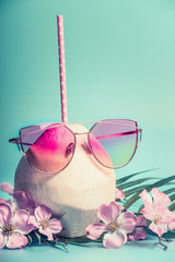 Wall Mural - Fresh coconut cocktail  with pink drink straw, sun glasses palm leaves and tropical flowers at blue turquoise background, front view. Tropical vacation travel concept