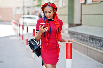 Wall Mural - Cute and slim african american girl in red dress with dreadlocks and backpack posed outdoor and looking at mobile phone on street. Stylish black model.