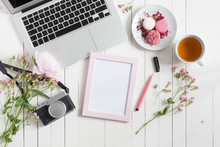 Flat Top View Of White Working Table With Gray Metal Laptop With Black Keyboard, Covered With Pink Flowers, With Camera, Pink Photo Frame And Felt Tip Pen For Sketching, Macarons And Tea Cup