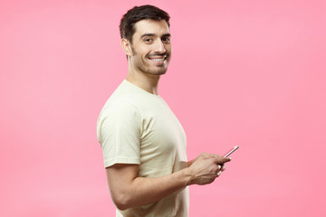 Side portrait of young handsome European man isolated on pink background dressed in casual T-shirt, holding cellphone in hands and looking at camera while waiting for answer in social network