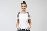 Indoor picture of young good-looking teenage girl isolated on gray background in white casual T-shirt, wearing glasses and gray backpack and jeans, feeling relaxed and positive, willing to walk