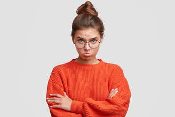 Upset beautiful female with offended look, curves lower lip in dissatisfaction, keeps hands crossed, wears oversized knitted sweater, being abused by close person. People and negative emotions