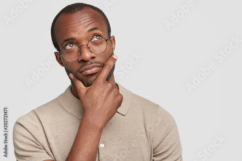 Fotografía Portrait of thoughtful dark skinned enterpreneur being deep in thoughts, contemplates about successful business project, holds chin, looks pensively upwards, isolated over white studio background