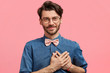 Photo of pleasant looking unshaven young male with pleased delighted look, keeps hands on heart, expresses love and devotion to girlfriend, isolated over pink background. People and emotions concept