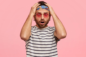 Candid shot of surprised terrified hippie guy scratches head and looks with puzzled expression, forgets about meeting with friends, isolated over pink background. People and subculture concept