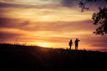 A Couple Of Man And Woman Is Walking And Looking At Amazing And Beautiful Sunset, With Tree On One Side, Silhouette
